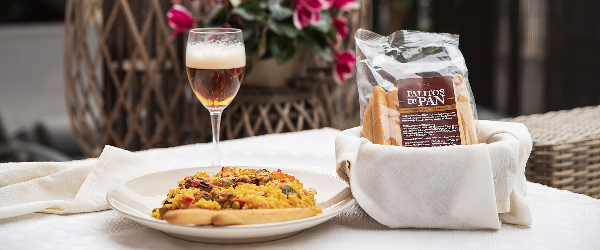 Foto de una mesa con una bolsa de palitos de pan, un plato de paella y una copa de cerveza